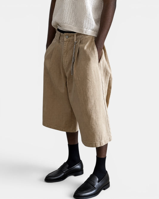 Nameless beige three-dimensional tailored wide-leg shorts with a silver chain detail. Model wears a white knit vest and black loafers.